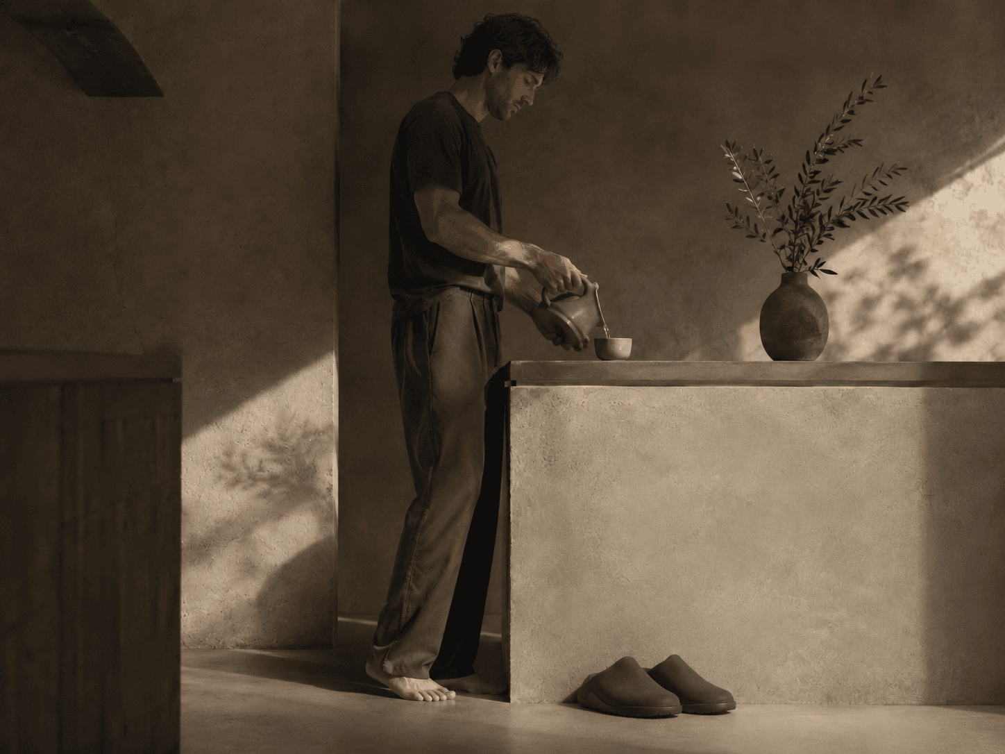 A man standing barefoot at a stone counter, pouring tea, with a pair of olive Inyo mules set on the floor beside him.