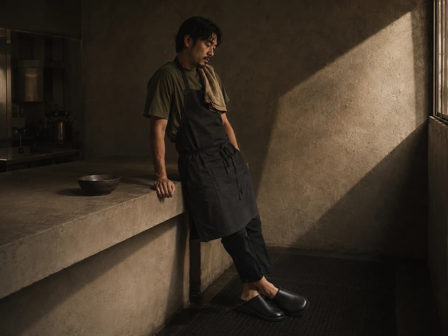 Chef in a black apron leaning against a concrete kitchen counter, weight shifted to one foot.