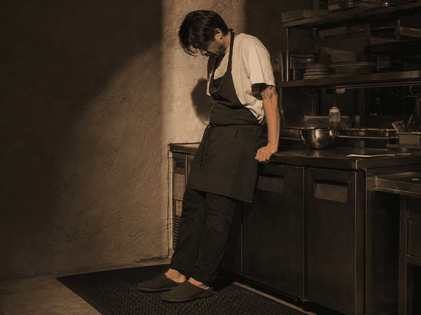 A kitchen worker in a white tee and dark apron leans back against a stainless-steel counter under warm directional light, wearing sage-green Inyo mules.